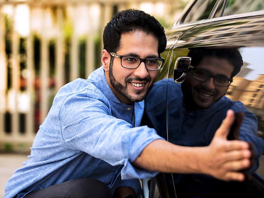 A man with black glasses is smiling and touching the shiny door of a black car next to him.