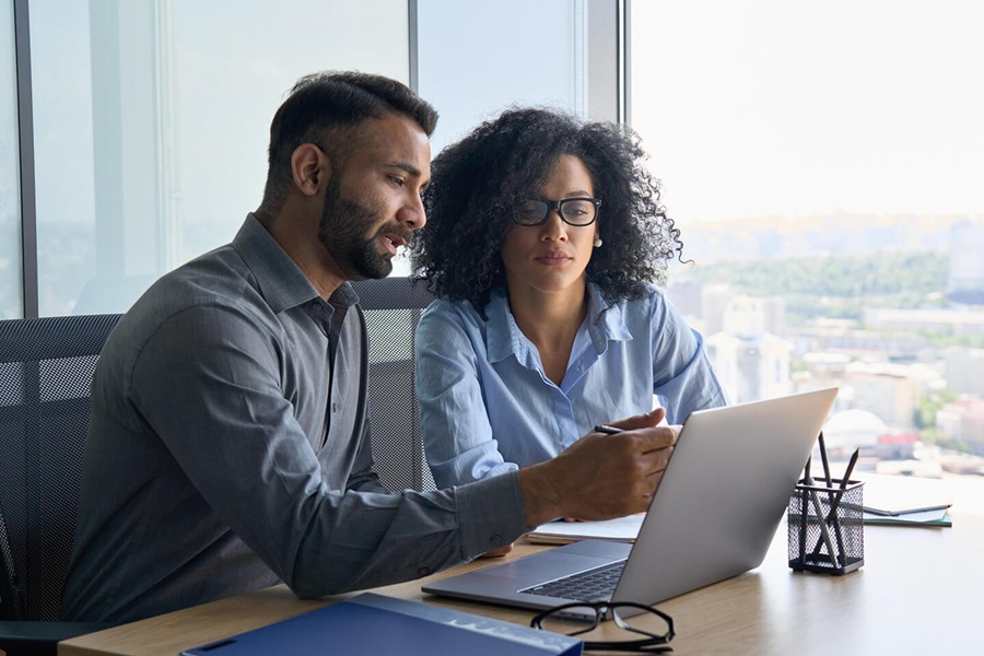 Two professionals reviewing work on a laptop by a window, man pointing at screen, woman looking on