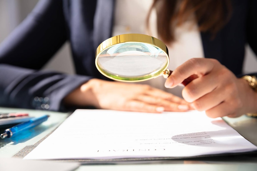 Person in business attire examining document with magnifying glass, papers visible on desk