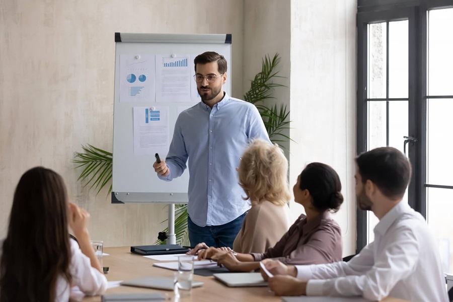 Man in light blue shirt presenting data charts on whiteboard to four attentive colleagues in meeting room