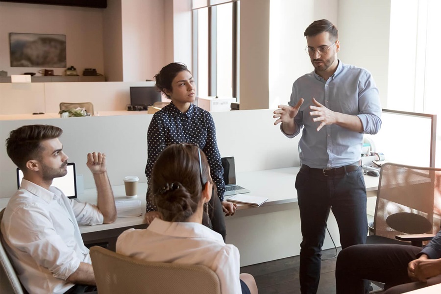Team meeting in modern office, standing man gesturing while explaining to three seated colleagues